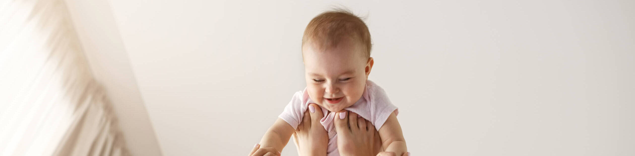 Young cheerful mother playing with her newborn baby lying on bed at home. Copy space.